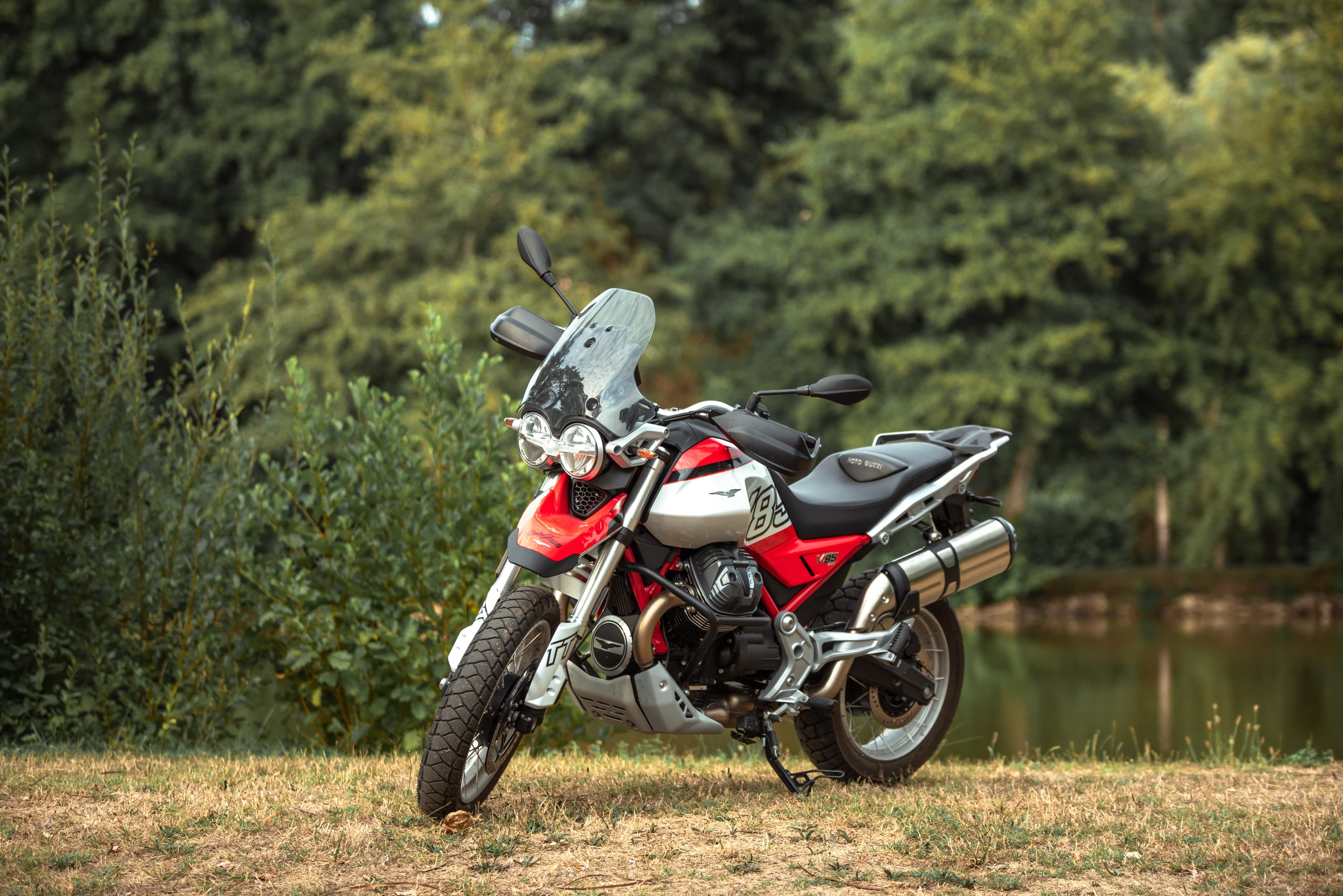 Moto Guzzi V85 TT rouge et blanche garée près d'un étang bordé d'arbres.